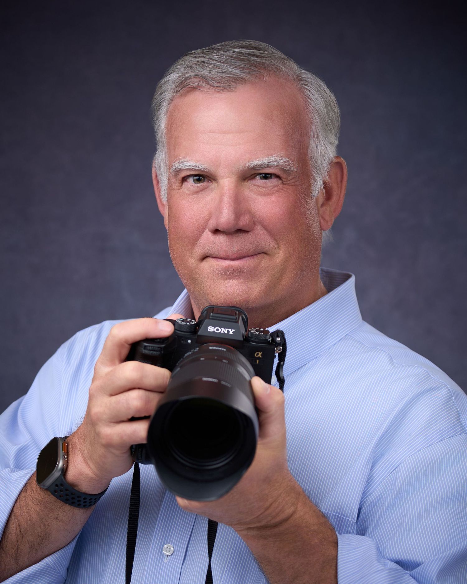 Middle-aged man with gray hair holding a Sony camera, wearing a light blue shirt, posed for a professional photographer portrait.
