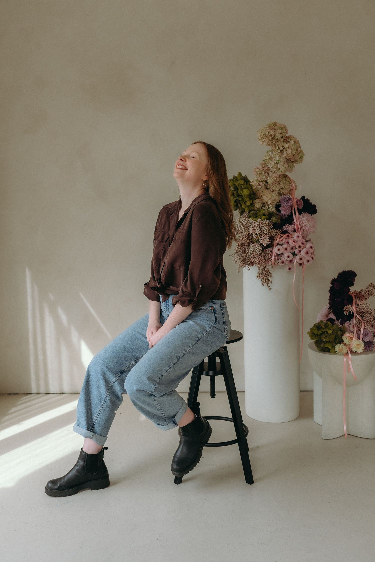 A person in a black button-down shirt and rolled-up jeans sits casually on a wooden chair against white curtains.