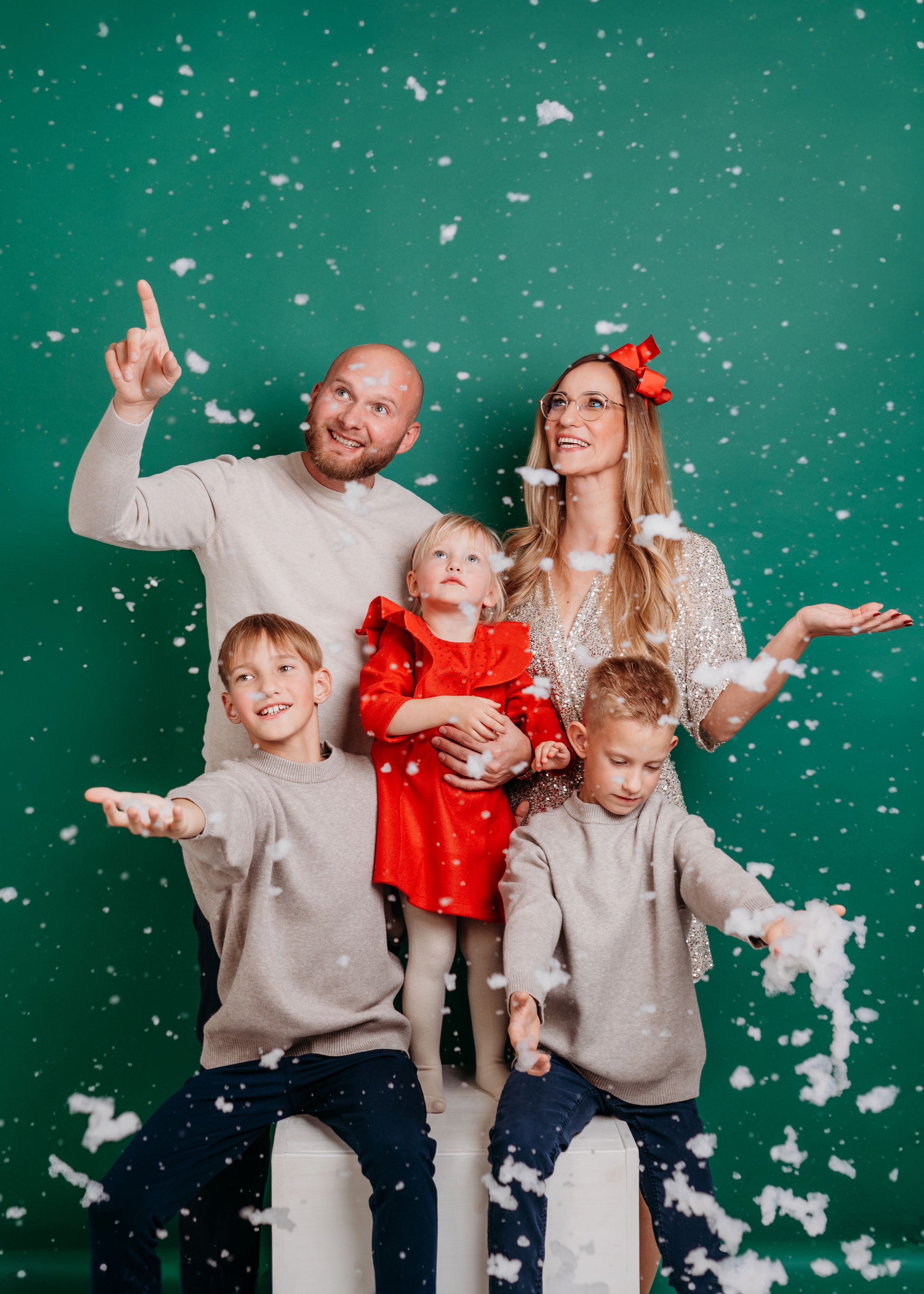 Family in matching beige sweaters playing with artificial snow against a green backdrop during a holiday photoshoot.