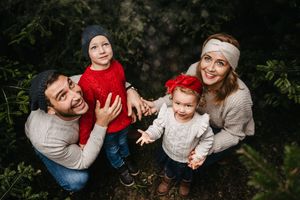 A family of four laughs together while posing outdoors against a dark green foliage background.