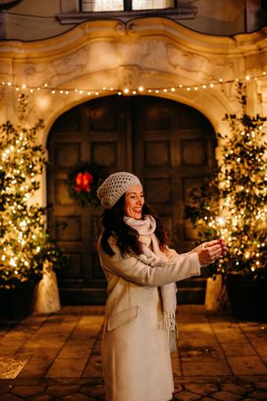 Person in white winter coat and knit hat dances joyfully among illuminated Christmas trees in stone courtyard at night.