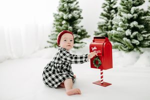 Small child in plaid dress sits in snowy winter scene with red Christmas mailbox and evergreen trees in background.