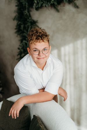 A child in a white button-down shirt poses on a gray couch with a warm smile and round glasses.