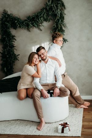 A family in white and beige outfits poses together on a white ottoman with evergreen garland decor and a gift box.