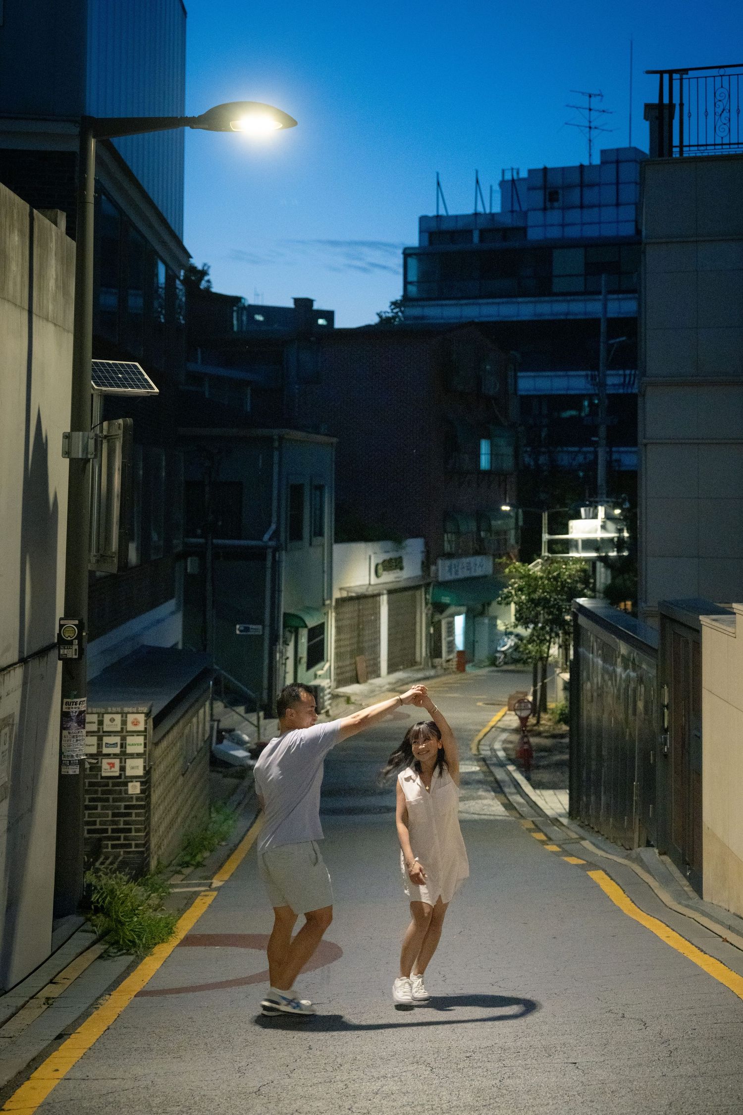 People in white outfits skateboarding down an urban alley at dusk with street lights illuminating the scene.