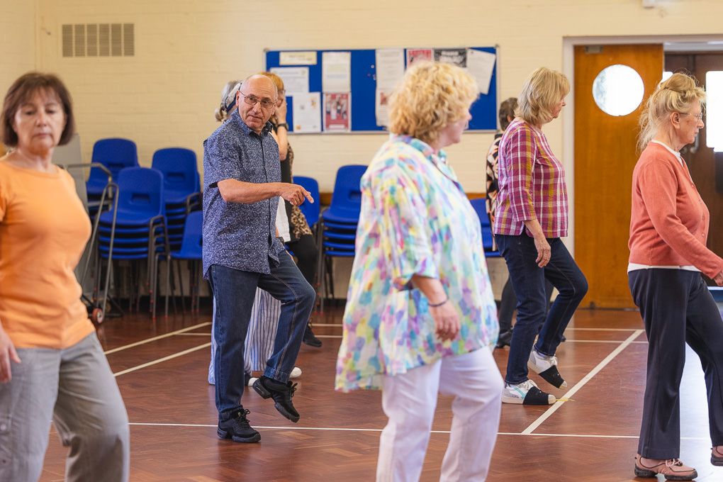 A group of seniors participating in a line dancing class at a community centre gymnasium in Hitchin.