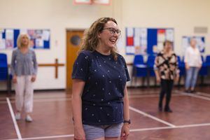 A person wearing glasses and a polka dot navy shirt laughs joyfully in a Letchworth community centre or gymnasium setting.