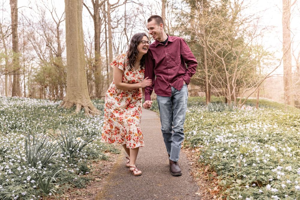A couple walks hand in hand through a wooded path with white spring flowers blooming along the ground.