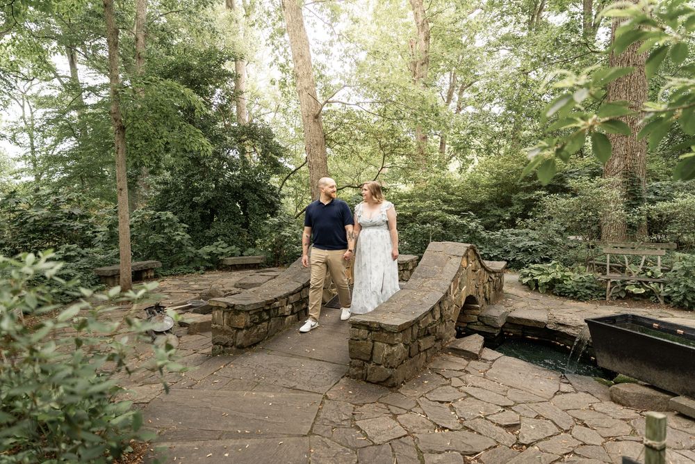 A couple stands on a rustic stone bridge surrounded by lush green forest during a summer photo session.