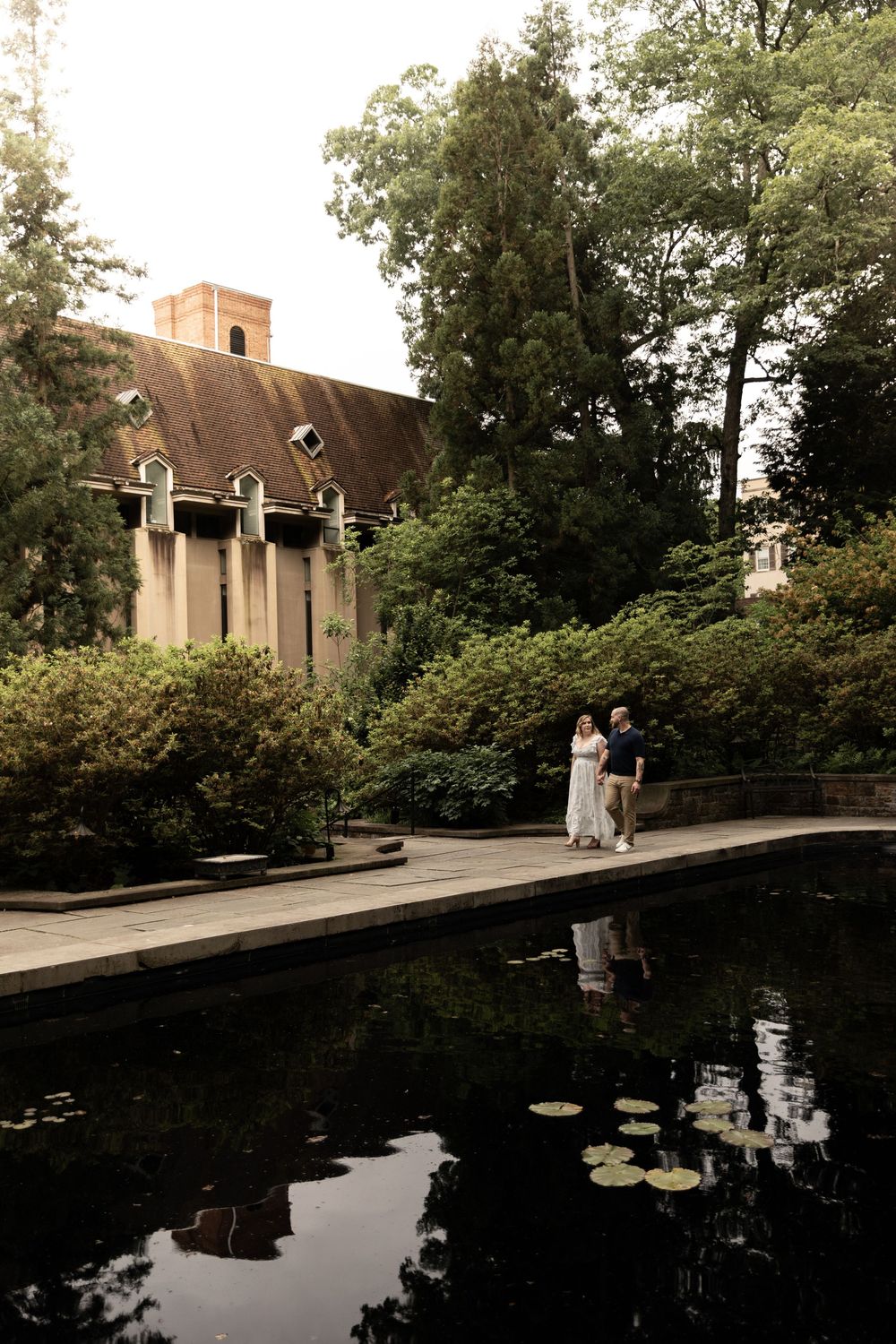 A serene pond with lily pads reflects trees and buildings on a tranquil day.