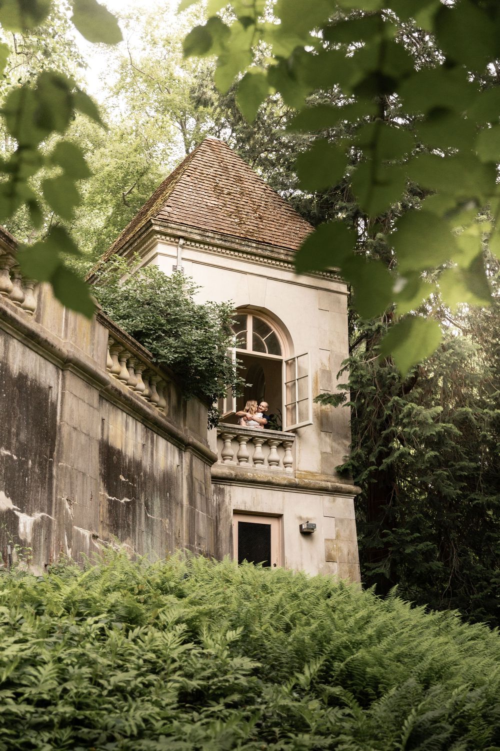 Old stone building with arched window peeking through lush green foliage and trees in a garden setting.