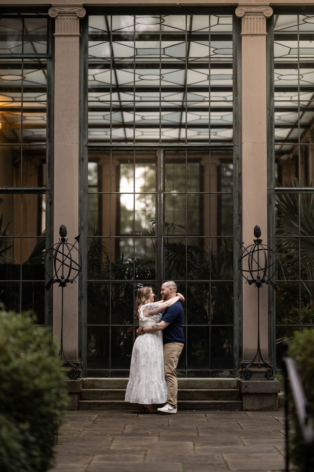 A romantic wedding couple shares a tender moment in front of a glass conservatory with elegant columns and iron details.
