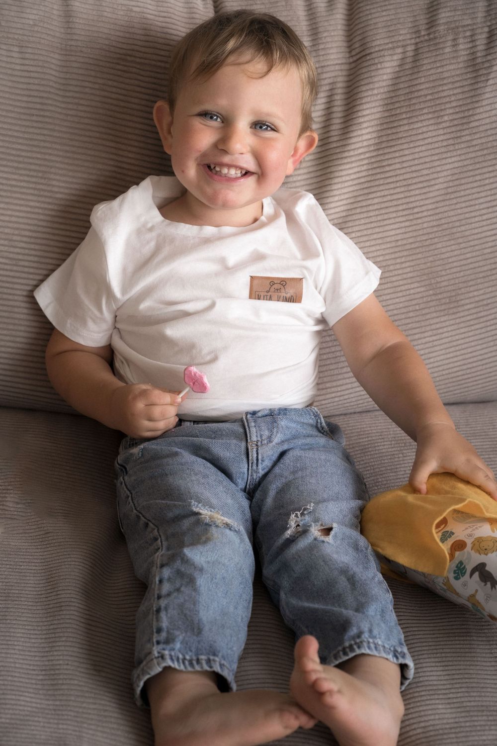 Kleinkind in weißem Hemd und Jeans sitzt lässig auf grauem Sofa.