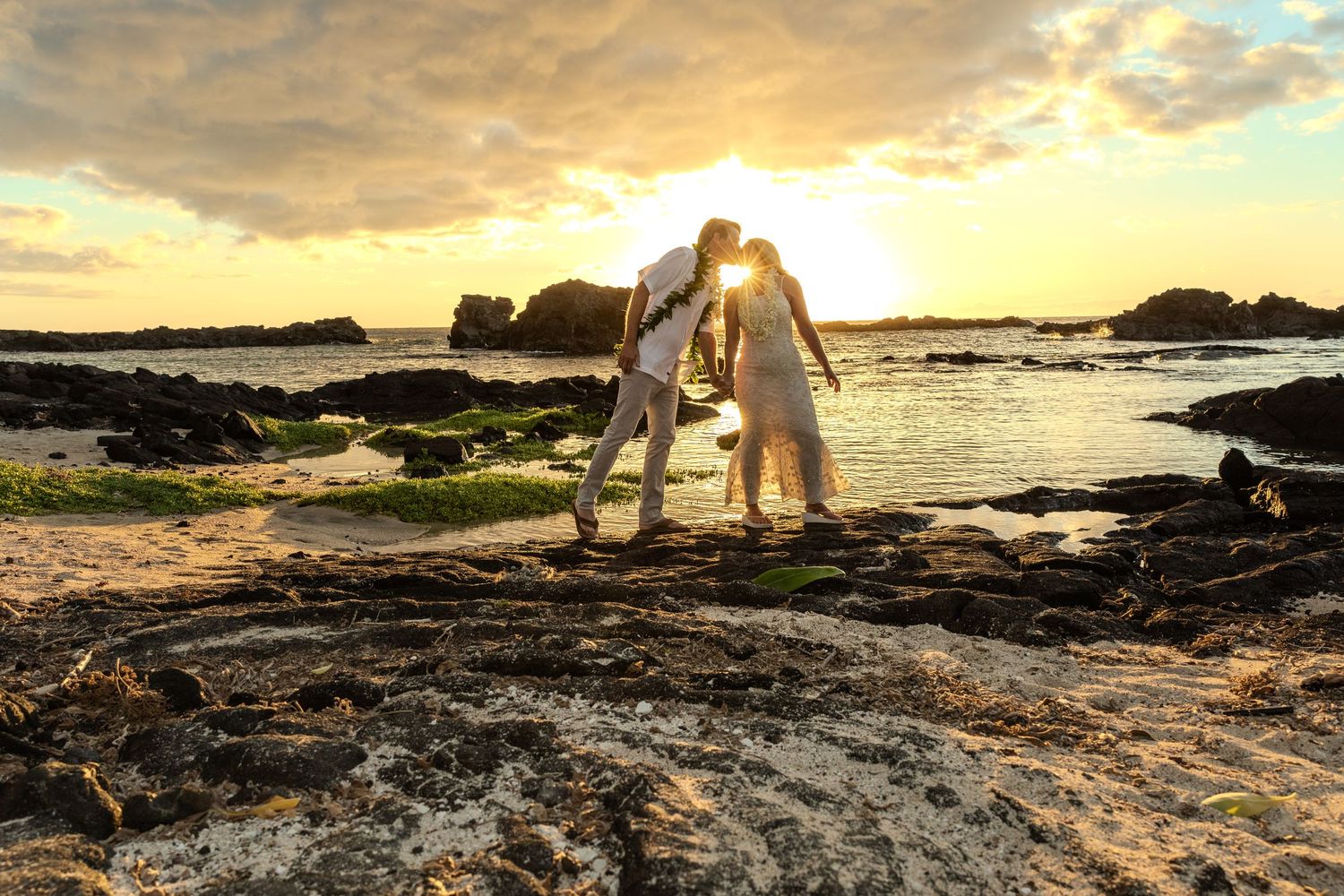 A romantic couple shares a tender moment on rocky beach terrain during a golden sunset.