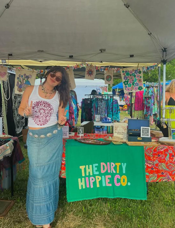 Colorful tie-dye clothing and merchandise displayed under vendor tents at an outdoor market with a green Dirty Hippie Co. banner.