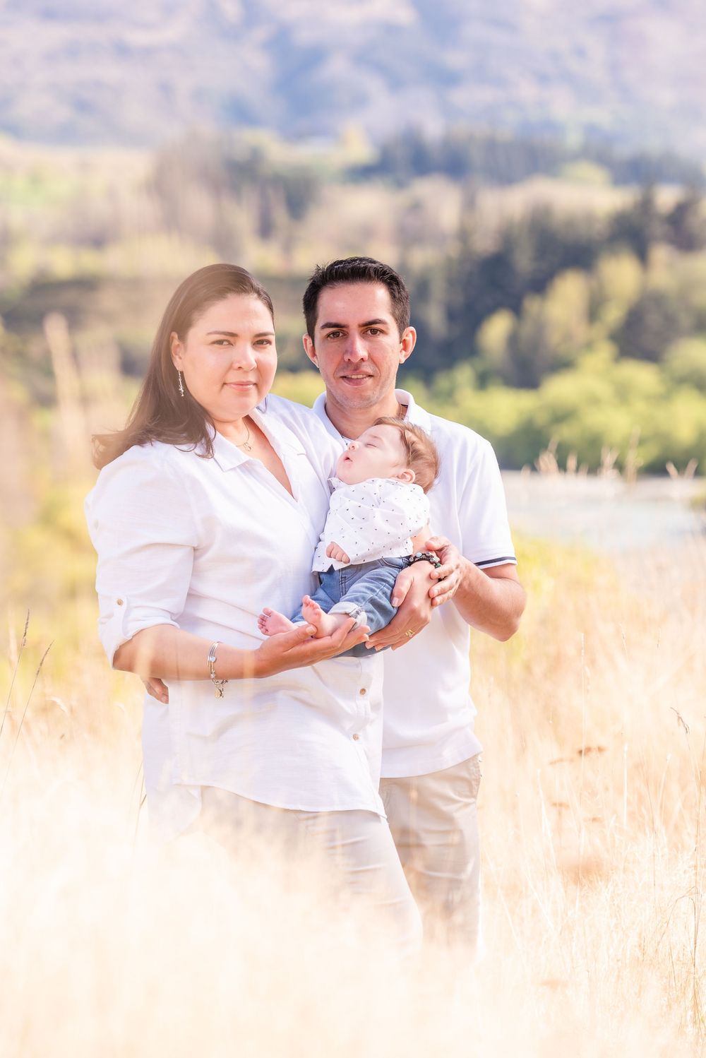 Young family in white shirts holding newborn baby in golden field with scenic mountain backdrop.