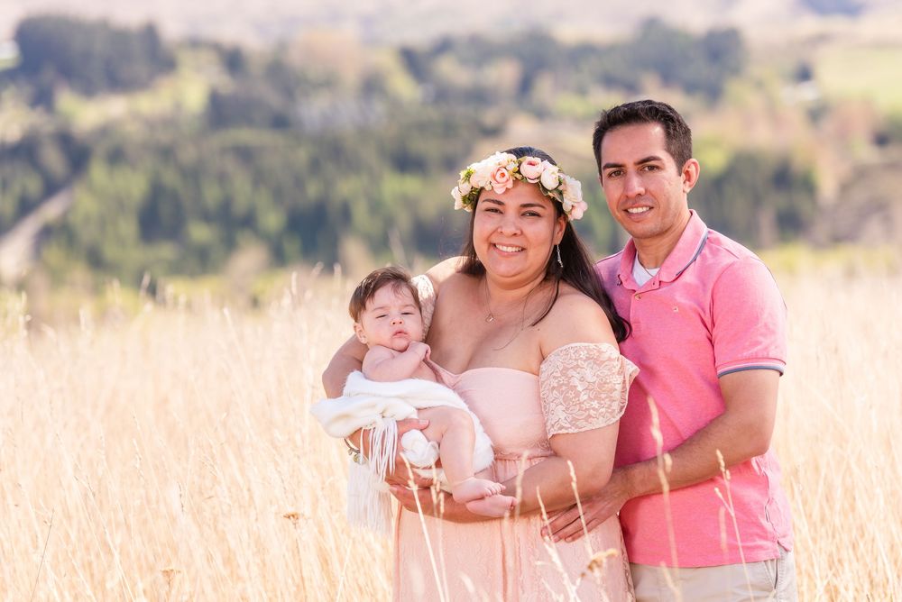 Family portrait of a couple holding their baby in a golden field with rolling hills and a floral crown.