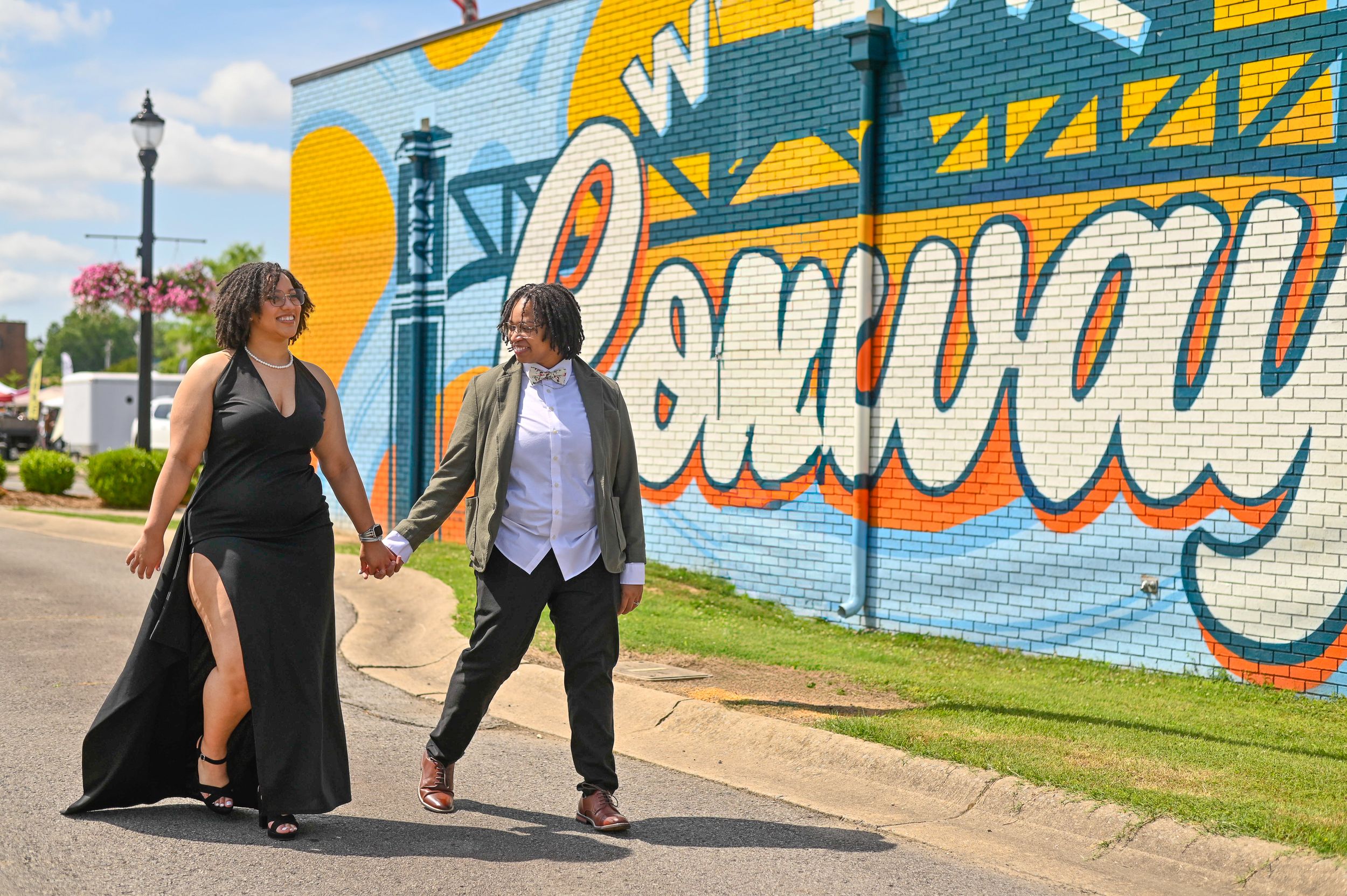 Two women holding hands walking past a colorful Conway mural on a sunny day.