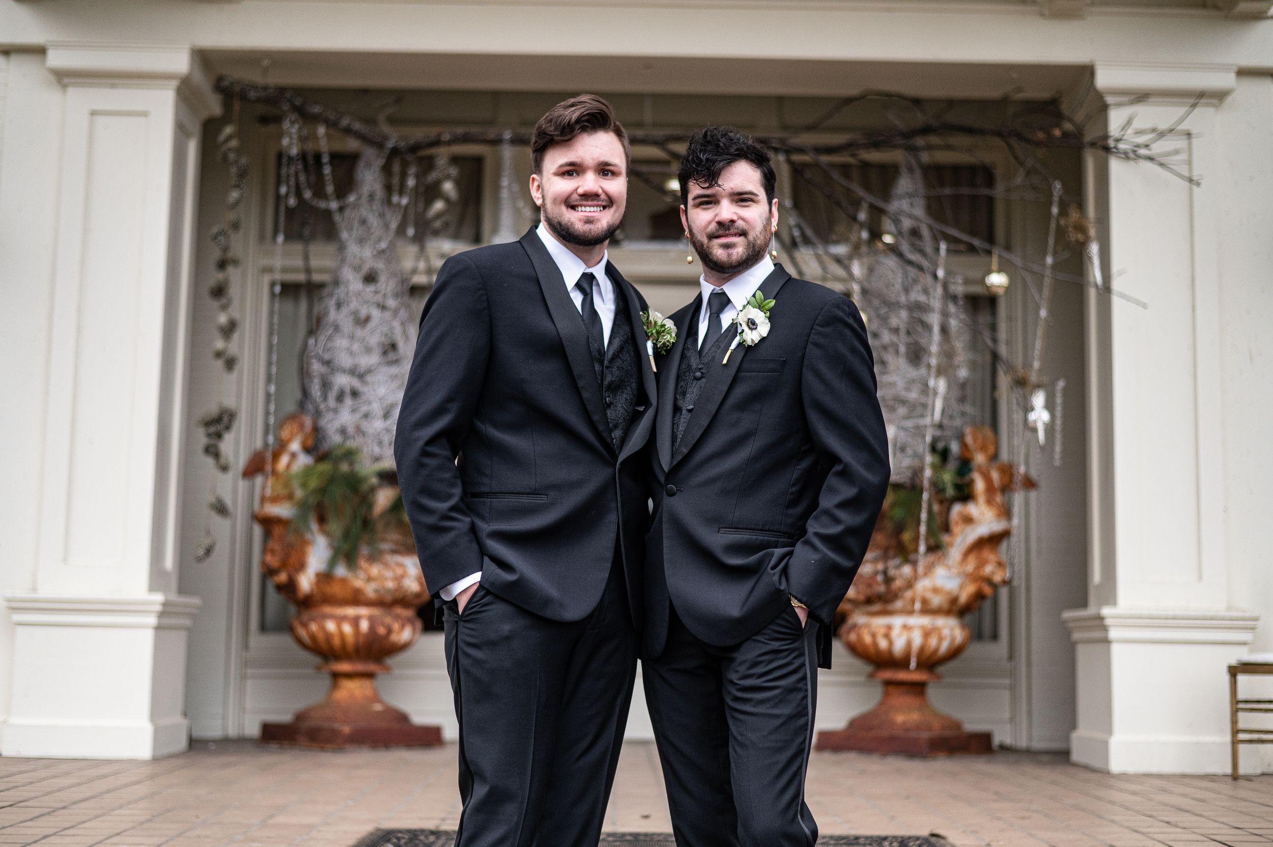 Two men in matching black suits with boutonnieres posing together outside a decorated venue.