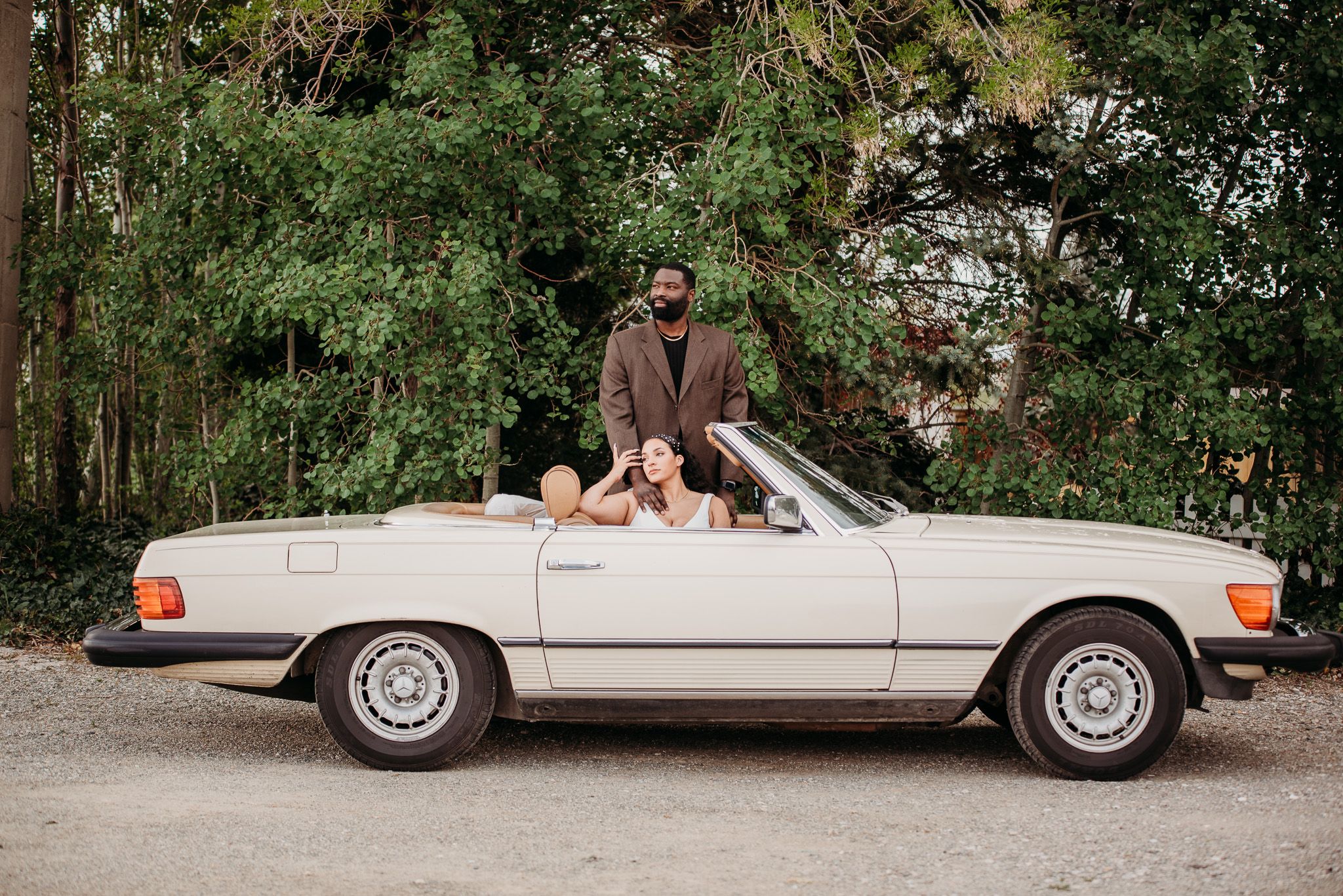 Couple posing with their classic white Mercedes convertible in a lush green setting.