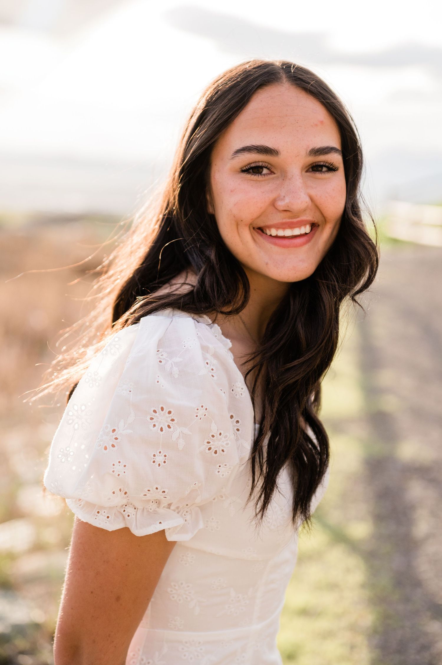 A portrait of someone in a white eyelet dress smiling in warm outdoor lighting.