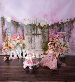 Pink and floral first birthday setup with rustic backdrop, string lights, and white 'ONE' letters on wooden floor.