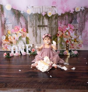 Pink cake smash photo session with floral decor and destroyed birthday cake on wooden floor.