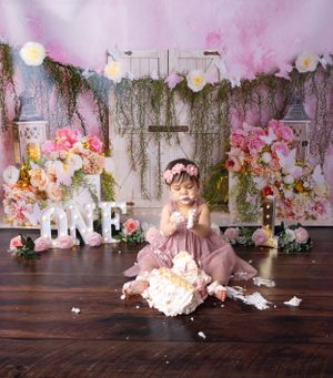 First birthday cake smash scene with pink dress and rustic floral backdrop setup.