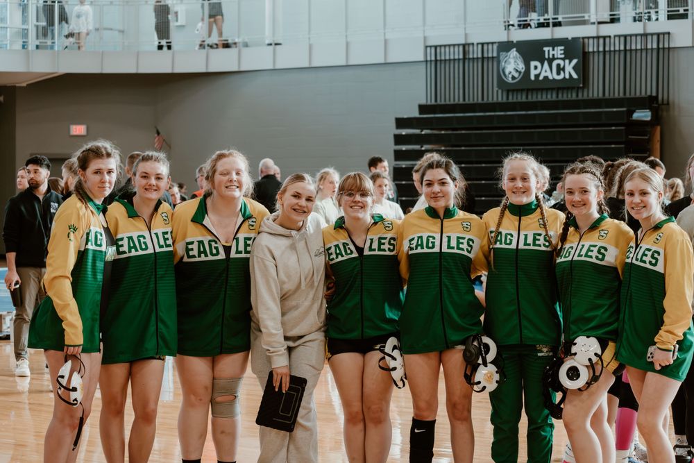 Girls wrestling squad in green and gold Eagles uniforms posing together inside a gymnasium.