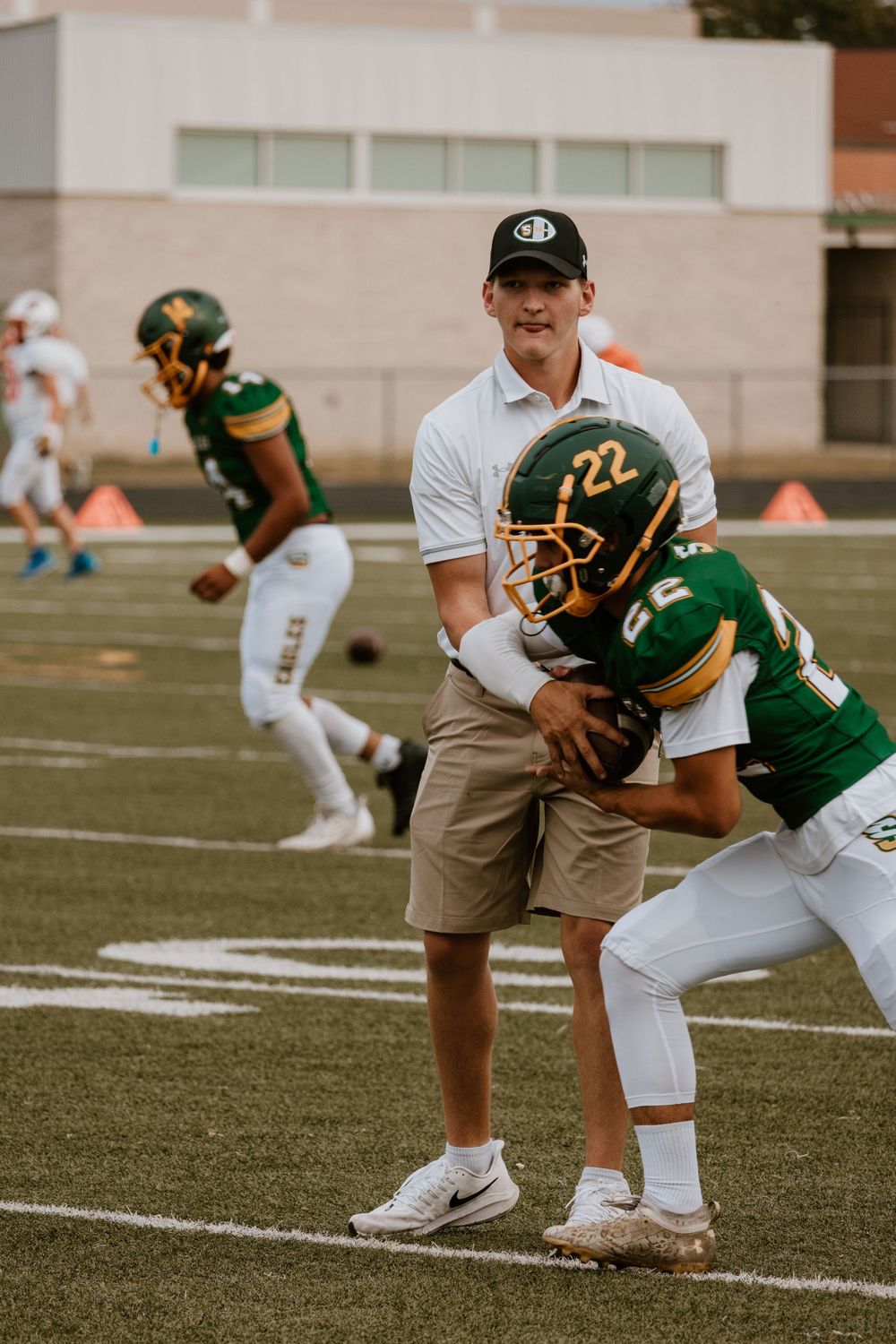 A football coach in white assists a young player wearing number 22 with technique during warm-ups on the field.