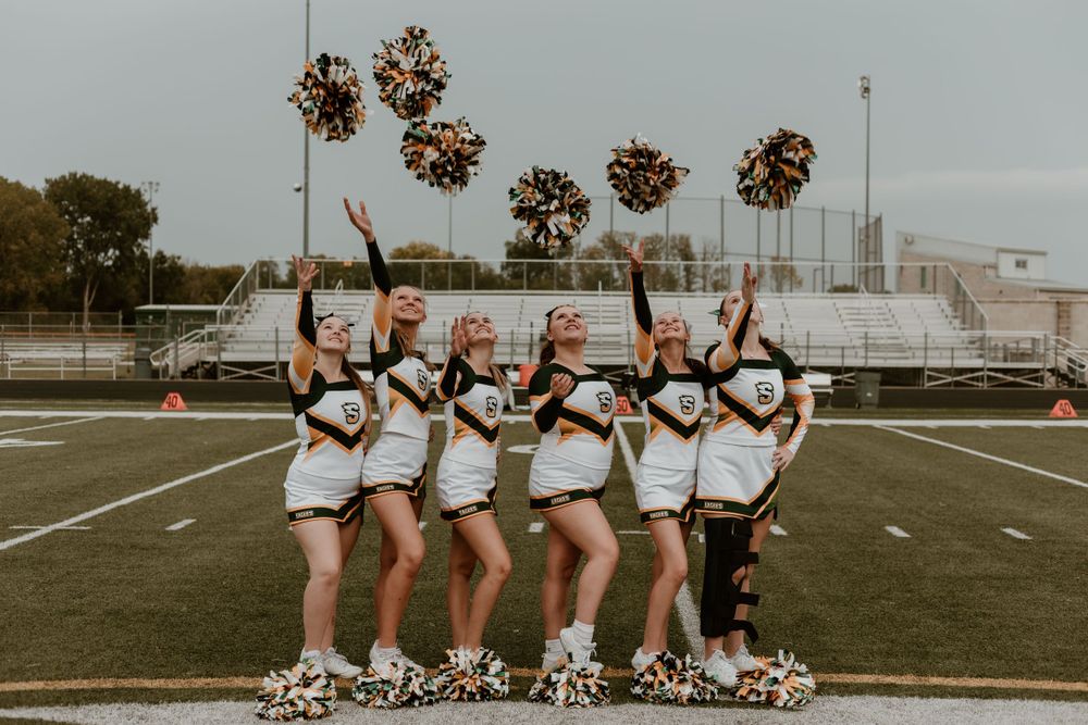 Five cheerleaders in white and gold uniforms pose in a line tossing pom-poms in the air on a football field.