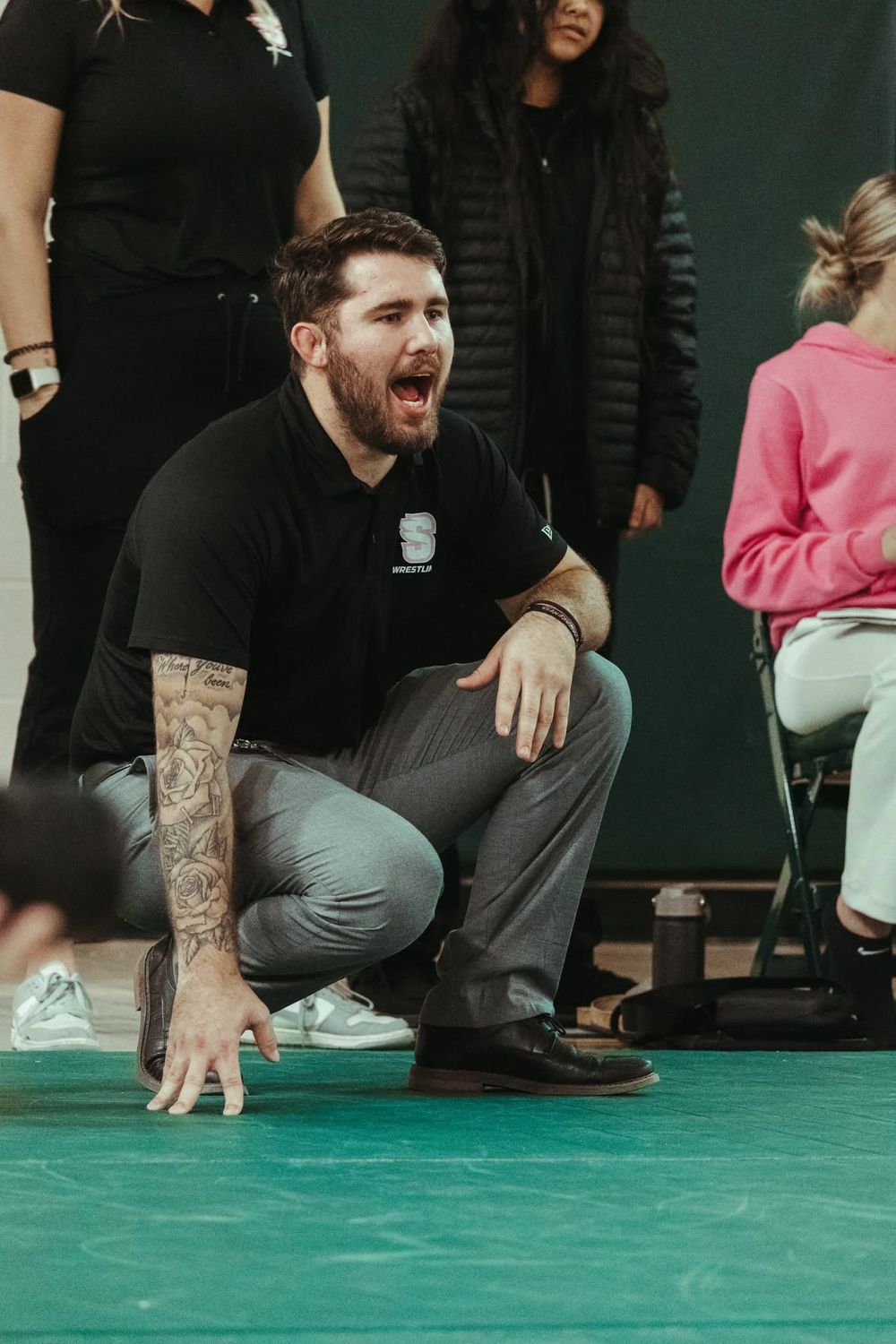 A tattooed male coach crouches and shouts encouragement during an indoor wrestling match on a green mat.