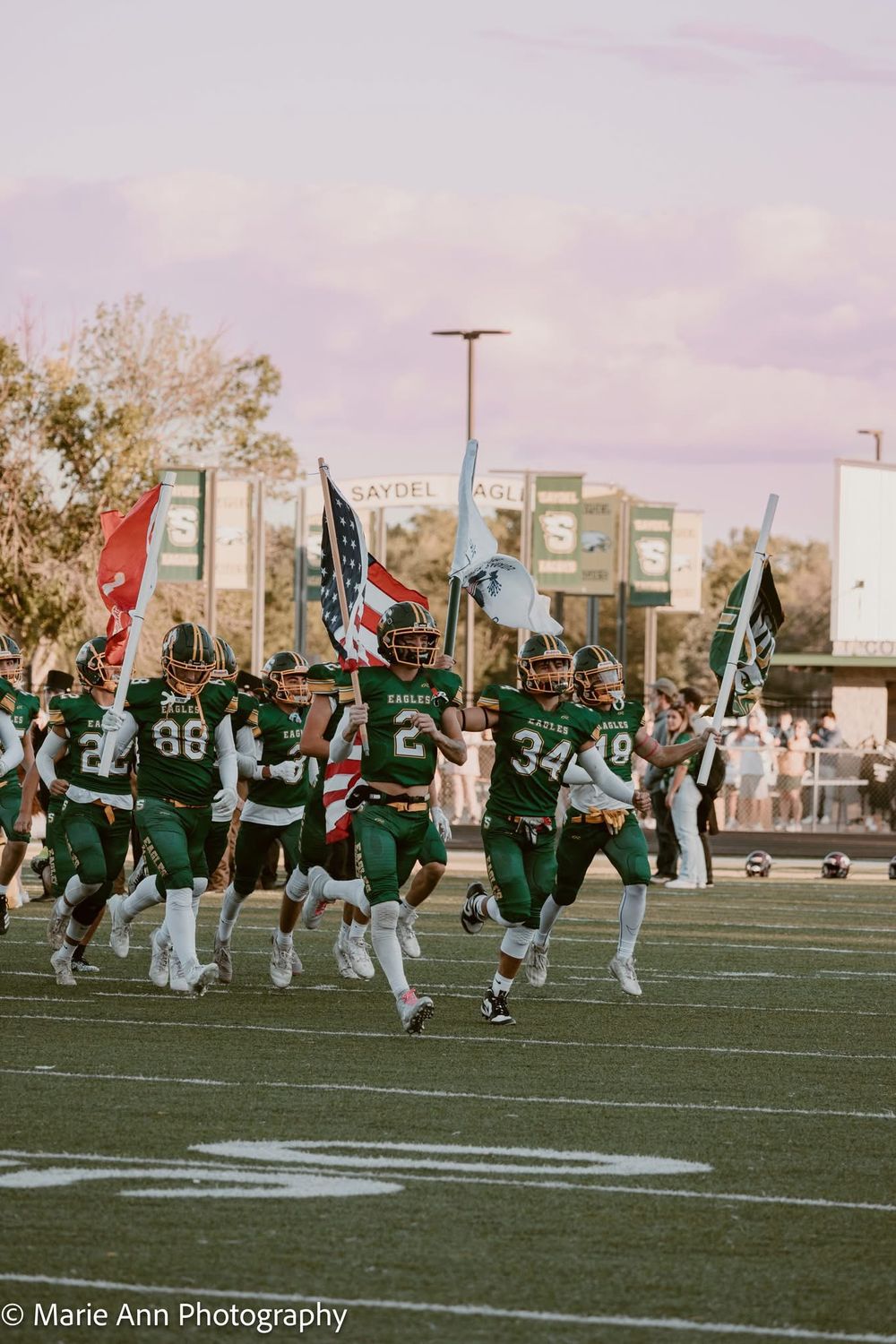 High school football players in green uniforms rush onto the field carrying American and team flags before a game.