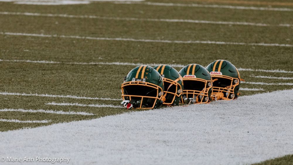 Three green and gold football helmets rest on a snow-dusted turf field during a cold weather game.