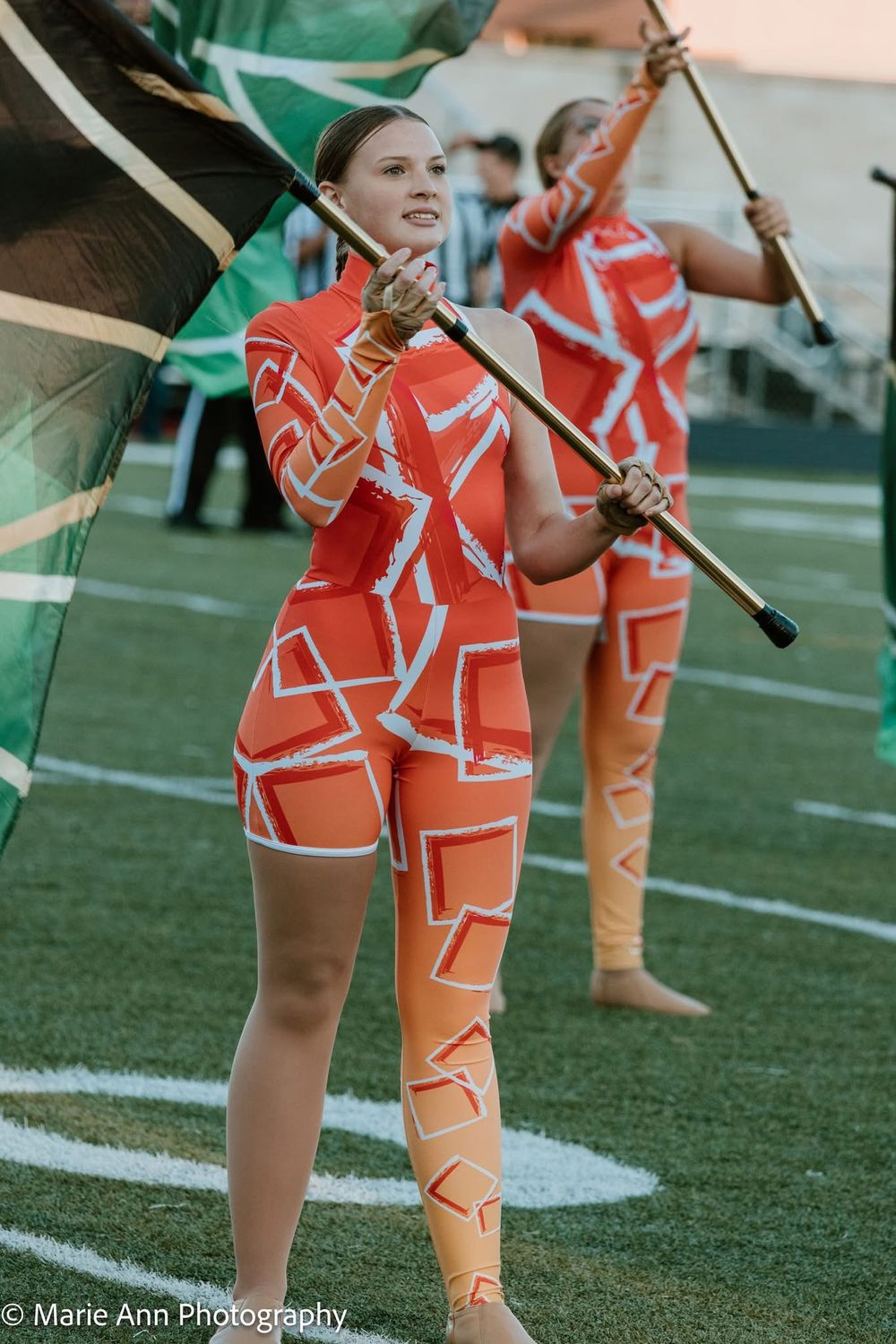 A color guard performer in an orange geometric costume twirls a flag during a marching band competition on a football field.