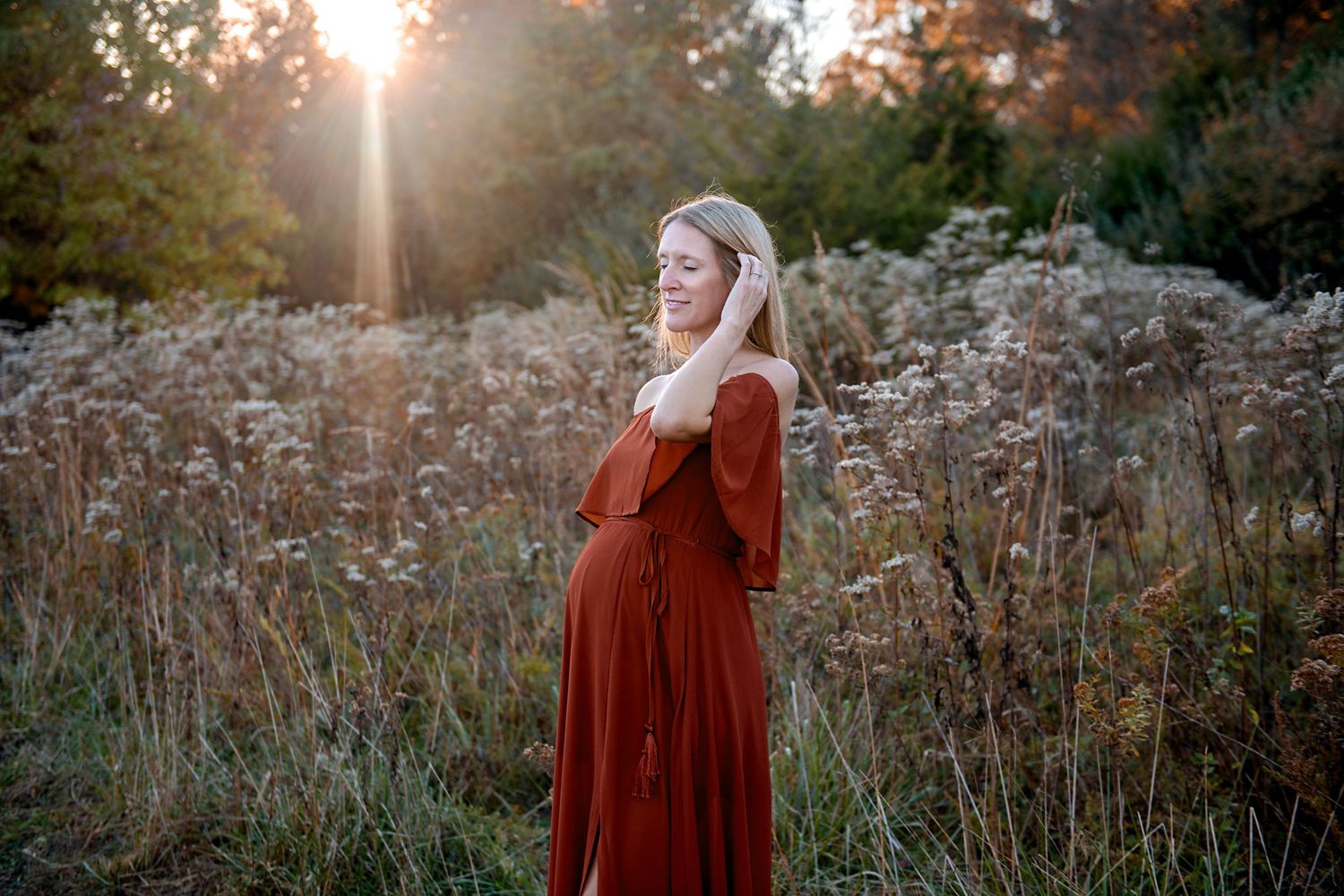 Woman in red dress poses in golden sunlit field during maternity photoshoot.