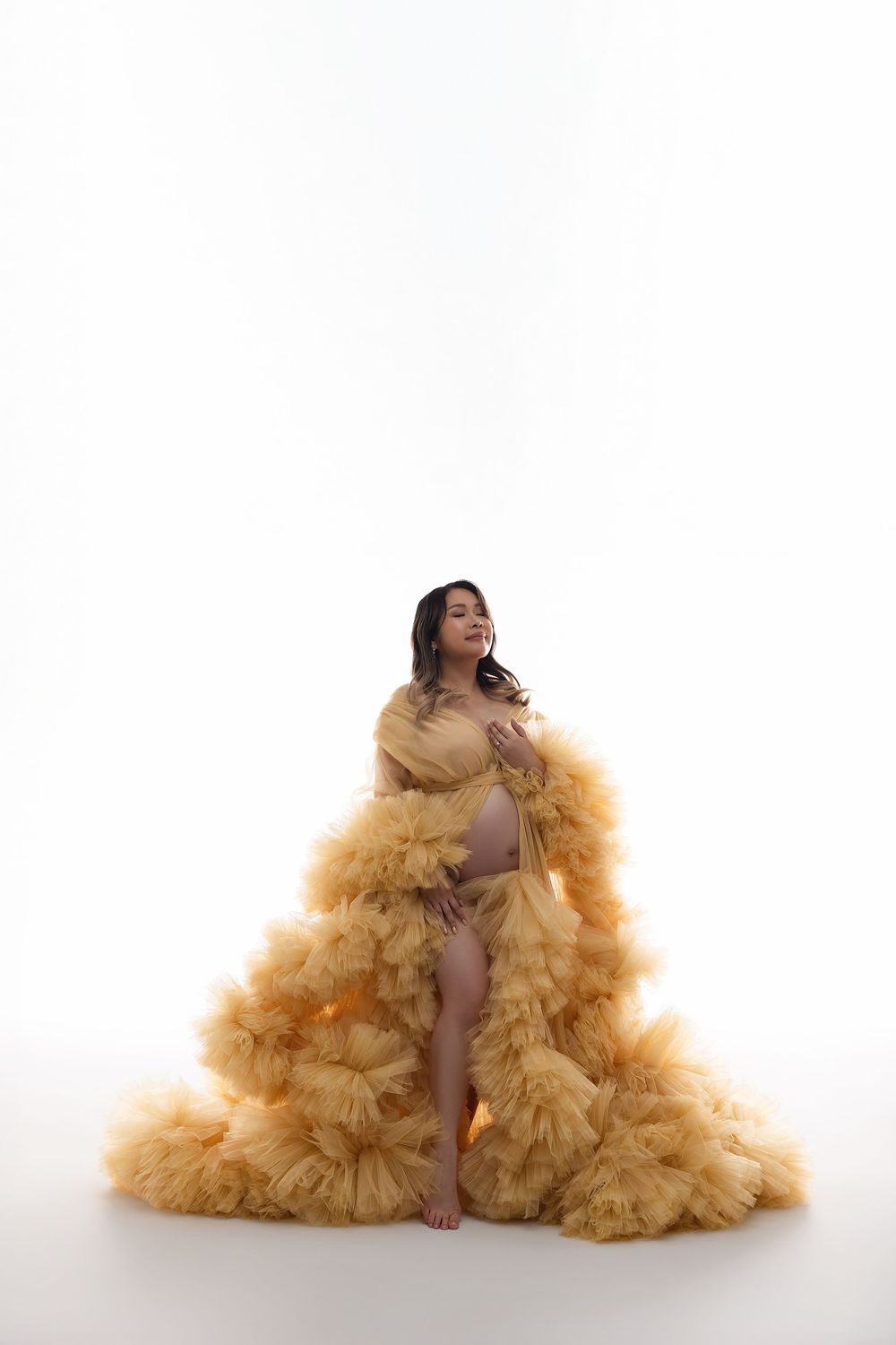 Pregnant woman in a flowing golden yellow tulle robe poses elegantly against a white background.
