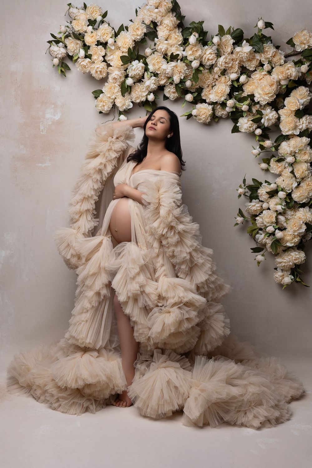 Artistic maternity photoshoot with cream tulle gown against floral backdrop of white roses in studio setting.