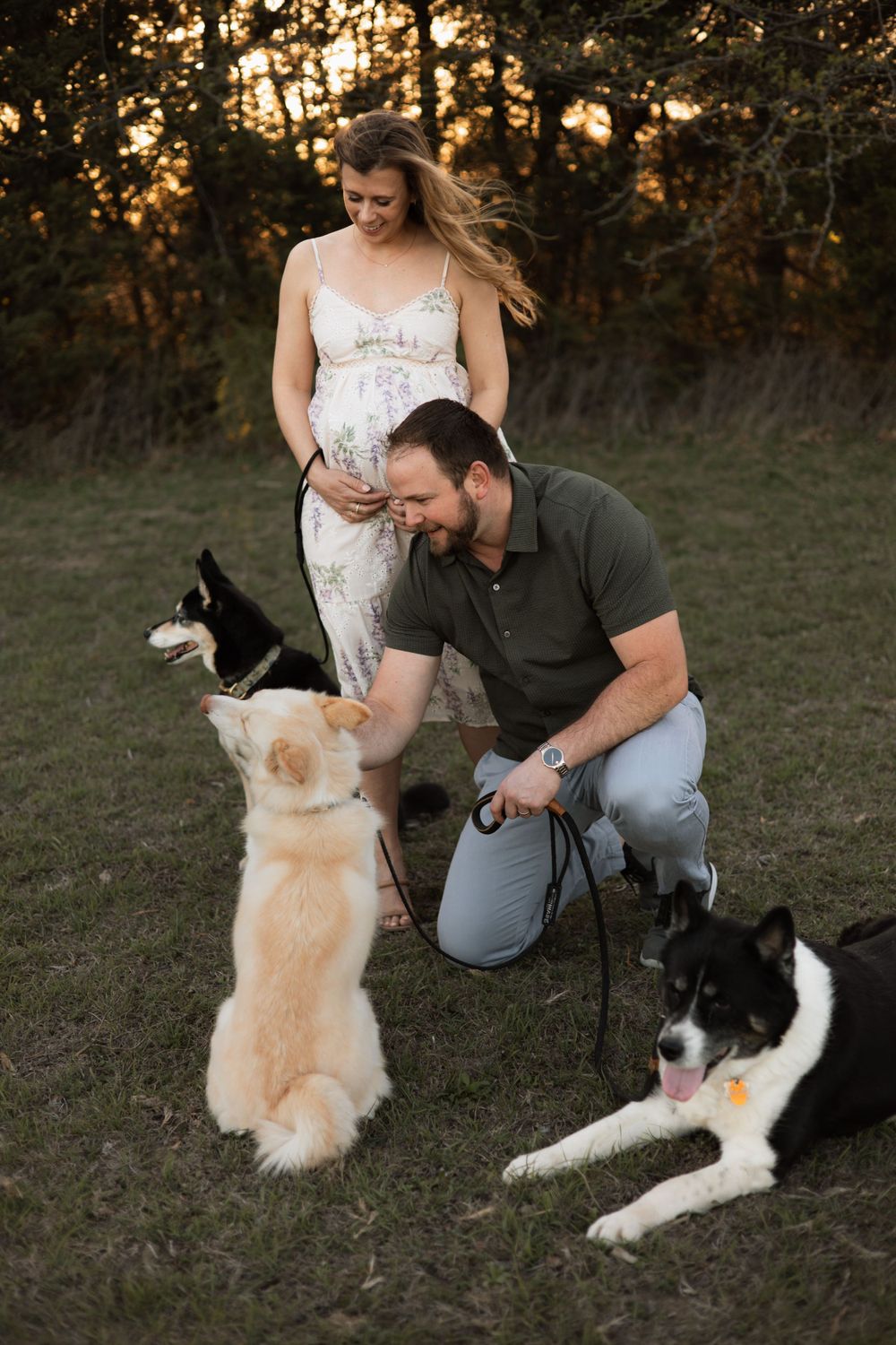 A couple and their two dogs share a tender moment at sunset in a grassy field.