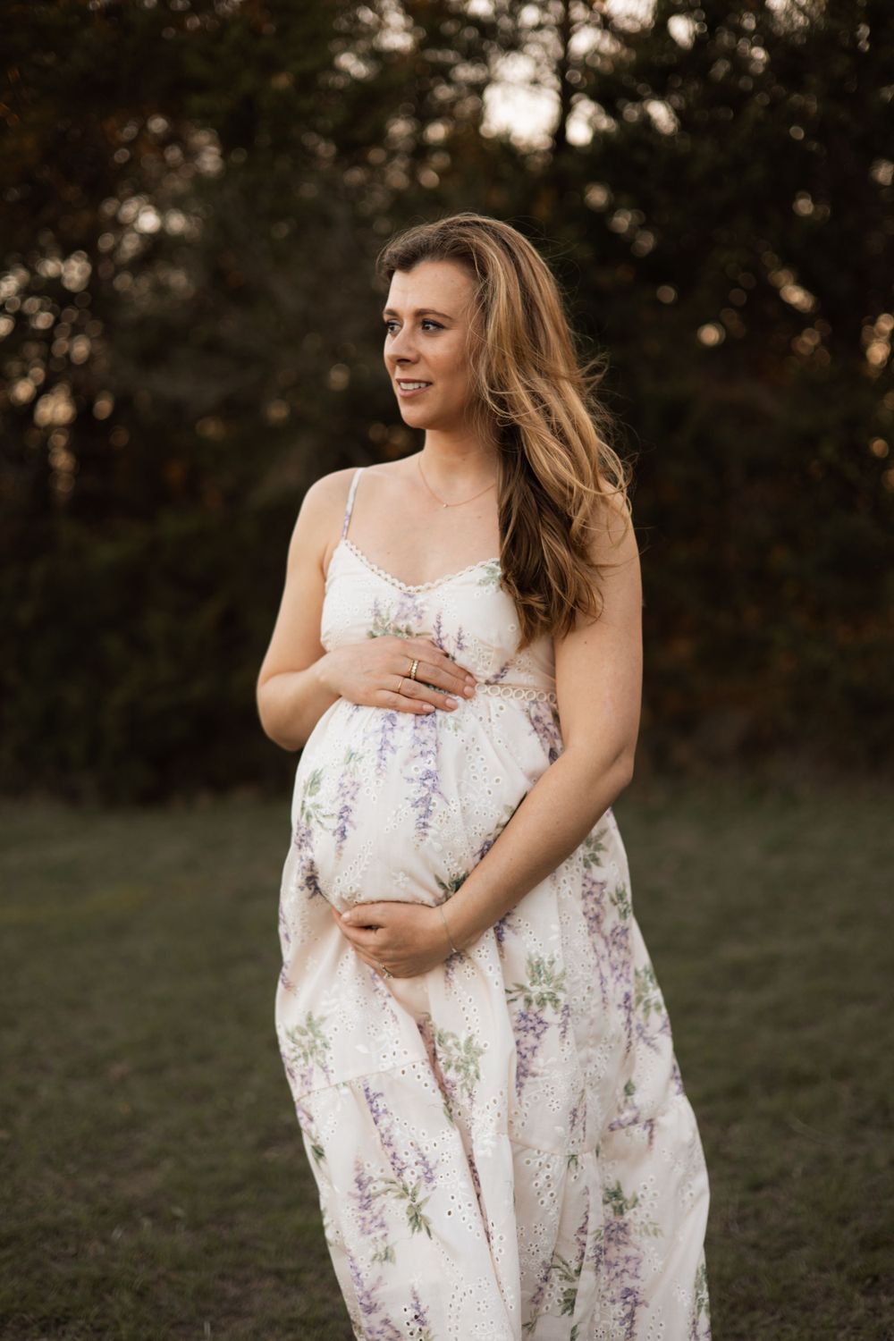 A pregnant woman in a floral white maxi dress poses in a field at sunset.