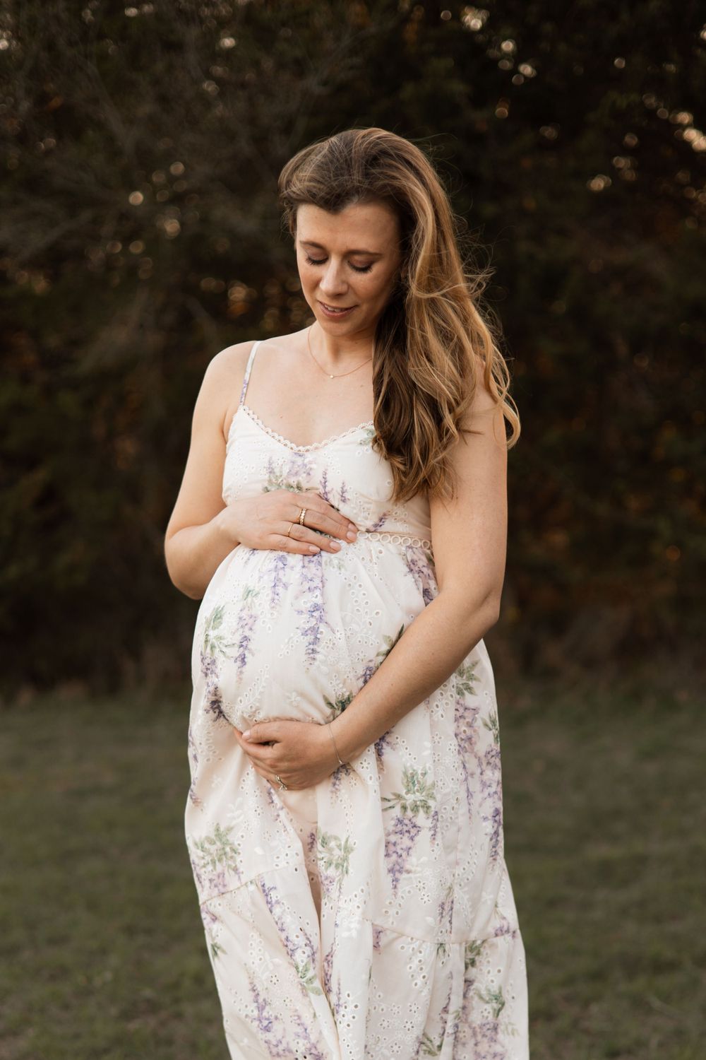 Person in a white floral dress cradles their pregnant belly while standing outdoors at sunset.