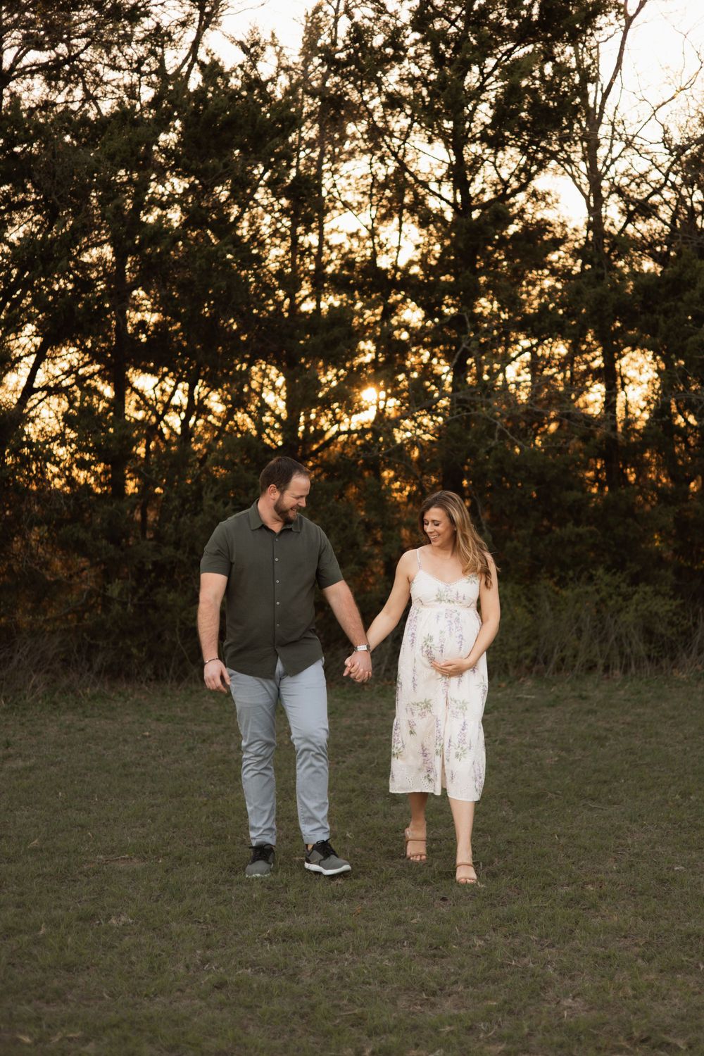 A couple walking hand in hand through a field at dusk with trees silhouetted in the background.