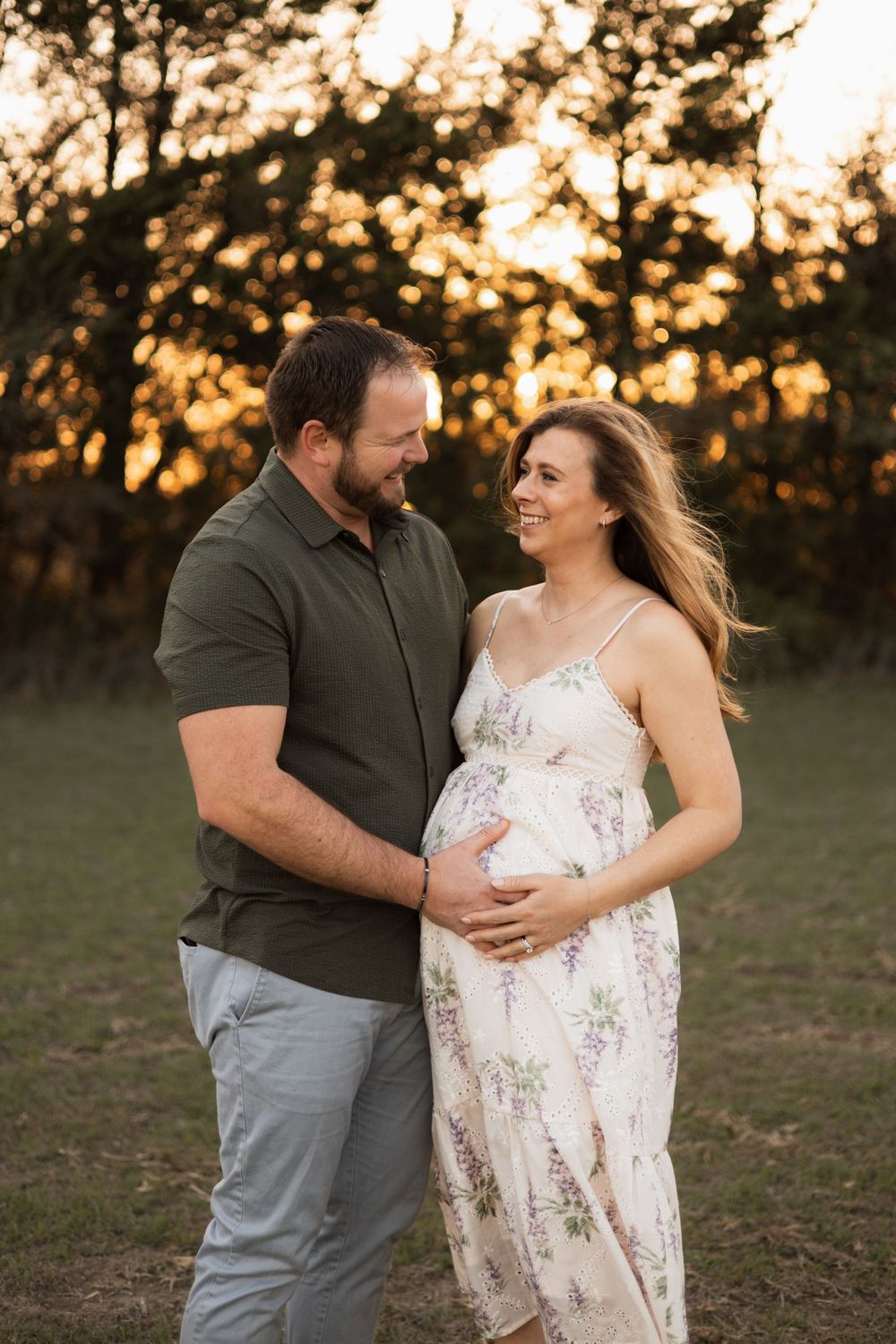 An expectant couple shares an intimate moment in a field during golden hour.