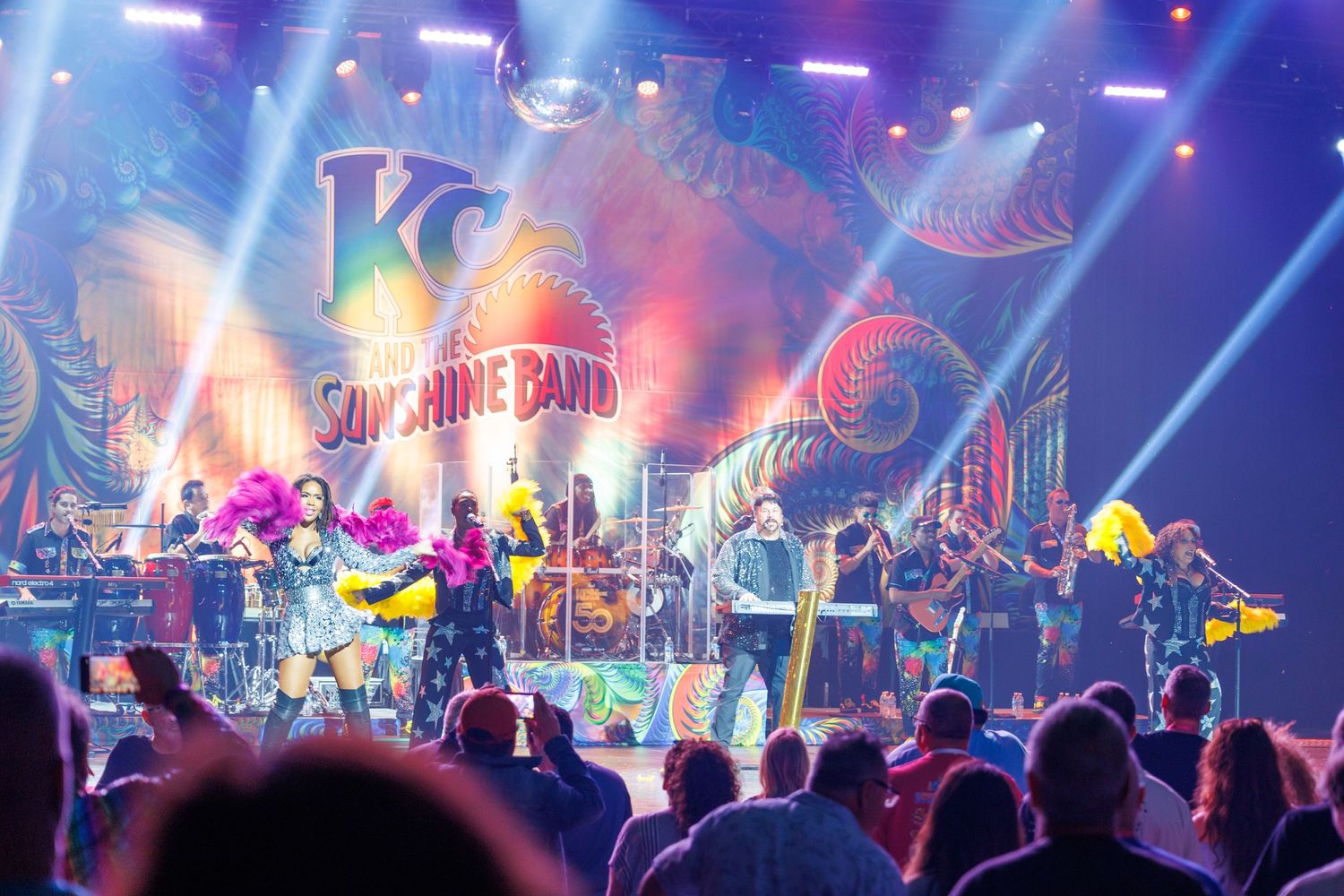 A vibrant concert performance with colorful stage lighting, musicians on stage, and a crowd watching from the audience floor.