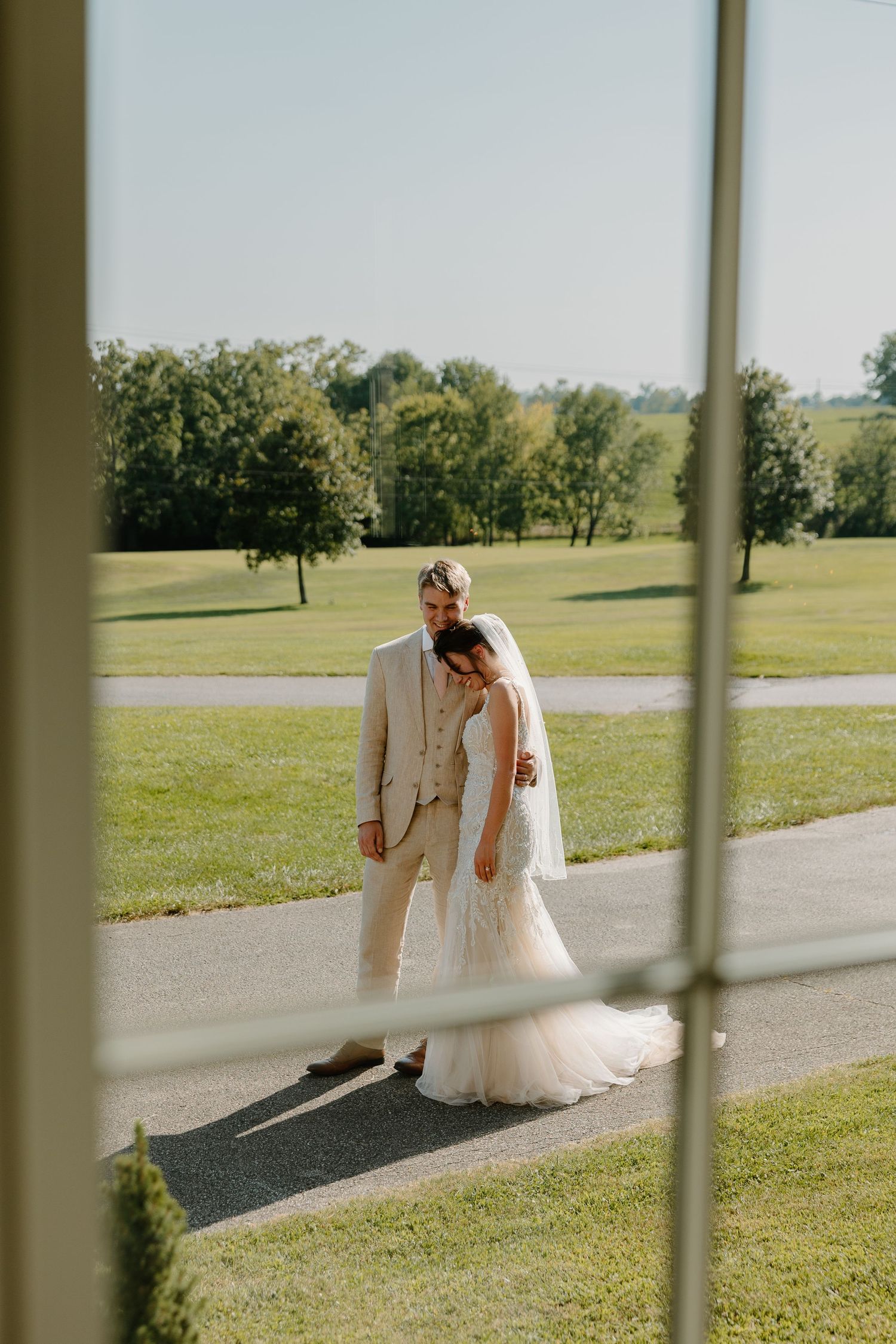 Wedding couple in light colored attire photographed through window panes on a sunny golf course.