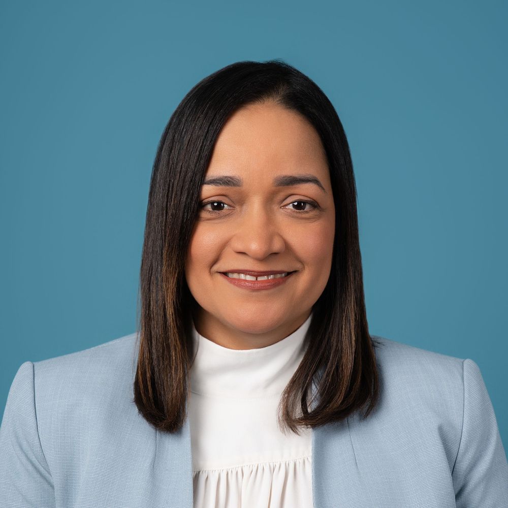 Professional headshots against a blue background showing different expressions and poses in a white turtleneck and gray blazer.