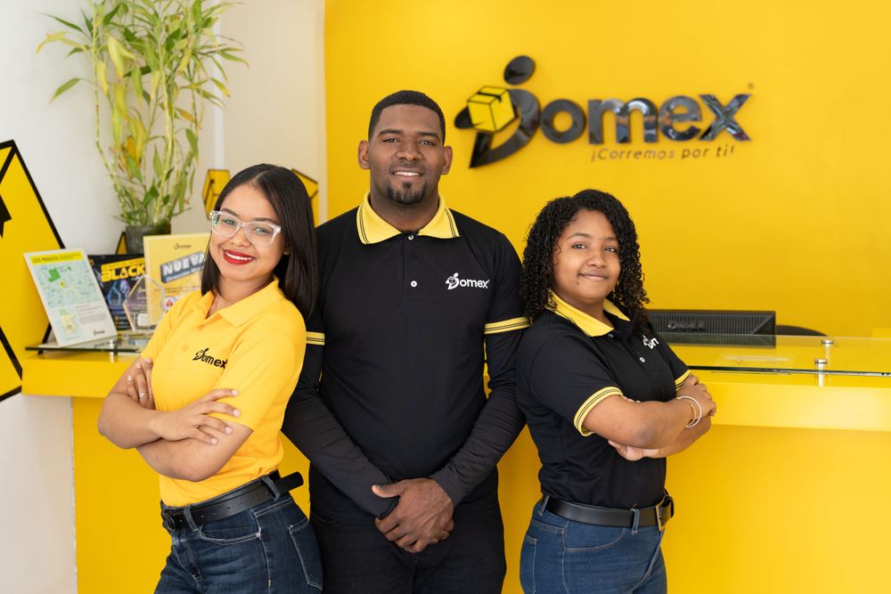 Three team members stand confidently in front of yellow Jomex company wall logo.