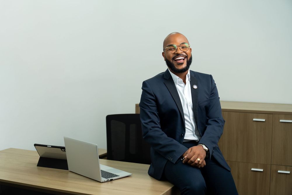 Business professional in navy suit smiling while sitting casually on office desk with laptop.