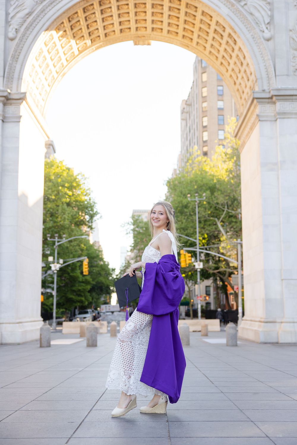 Graduate in lace dress poses with purple gown under Washington Square Arch.