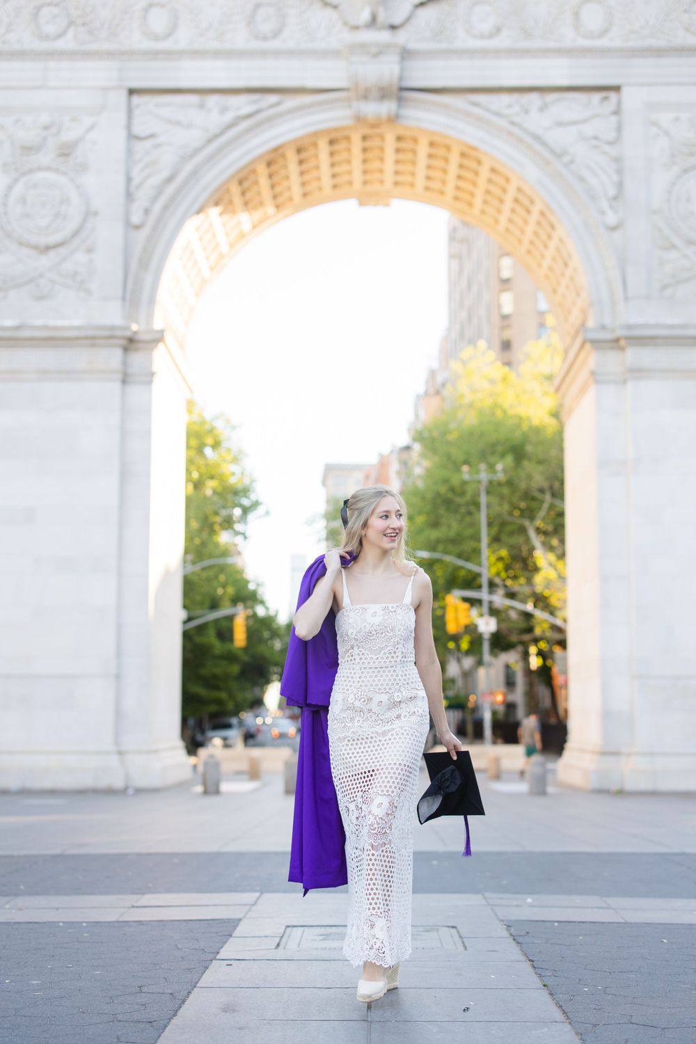 Graduate in white lace dress carries purple gown through Washington Square Arch.